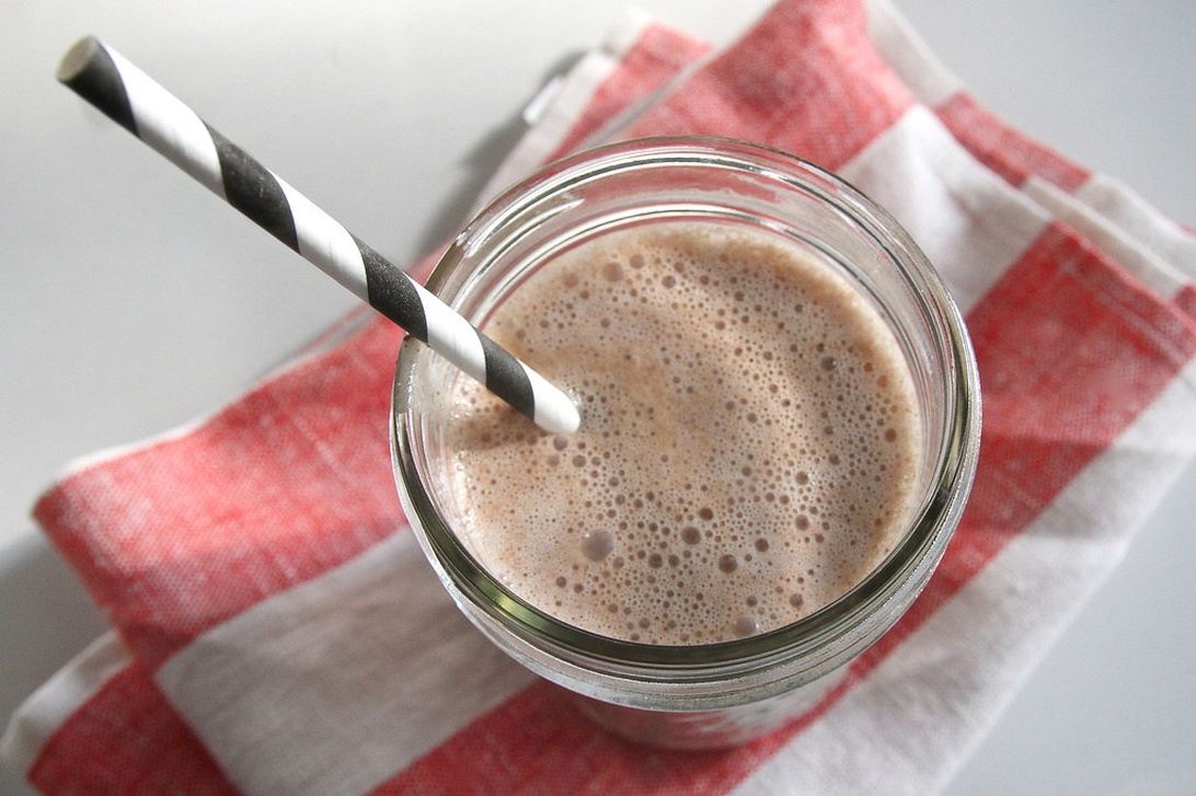 Ett glas med en återhämtningssmoothie eller proteinshake med ett svartvitt sugrör, placerat på en röd- och vitrutig duk, symboliserar näringsintag efter träning.